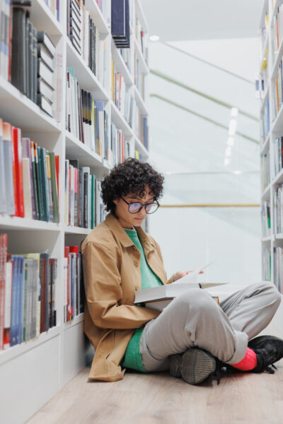 female student reads book sitting among the bookshelves in library or coworking.